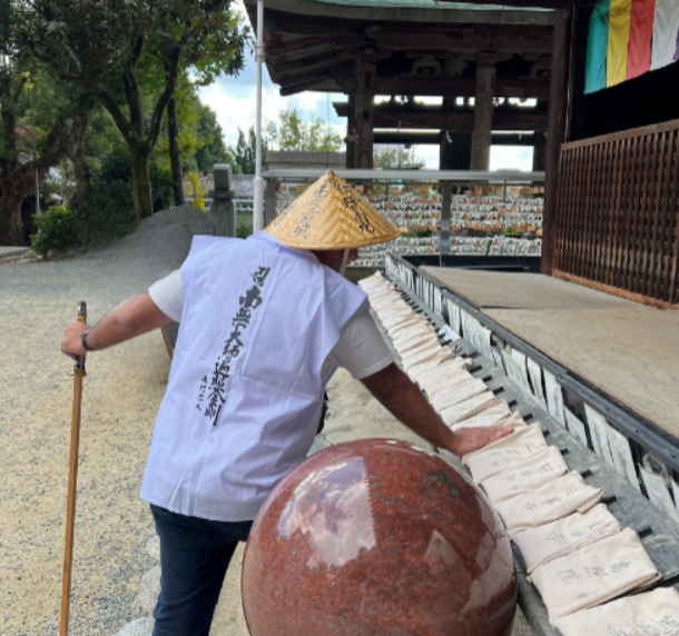 A white-clad pilgrim in a woven hat with a wooden staff at a temple hall, beside rows of sutra bags
